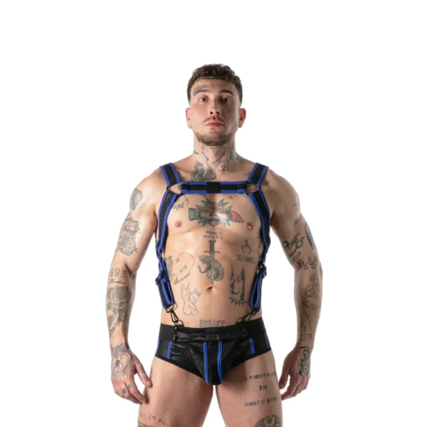 A tattooed person stands facing forwards, wearing a black and blue harness and matching underwear, against a plain white background.