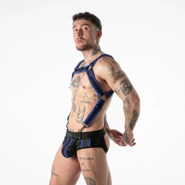 A tattooed person poses in a blue and black harness and matching underwear against a plain white background, looking over their shoulder with arms behind their back.