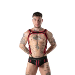 A muscular, tattooed man poses against a white background wearing a black and red harness and matching shorts, with his hands behind his back.
