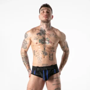 A muscular, tattooed man stands confidently, wearing black shorts with blue accents. He looks slightly upward with hands on his hips, set against a plain white background.