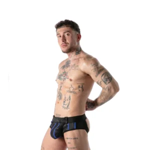 A tattooed man stands in profile against a white background, wearing black and blue harness-style underwear, with his hands resting on his hips and looking towards the camera.