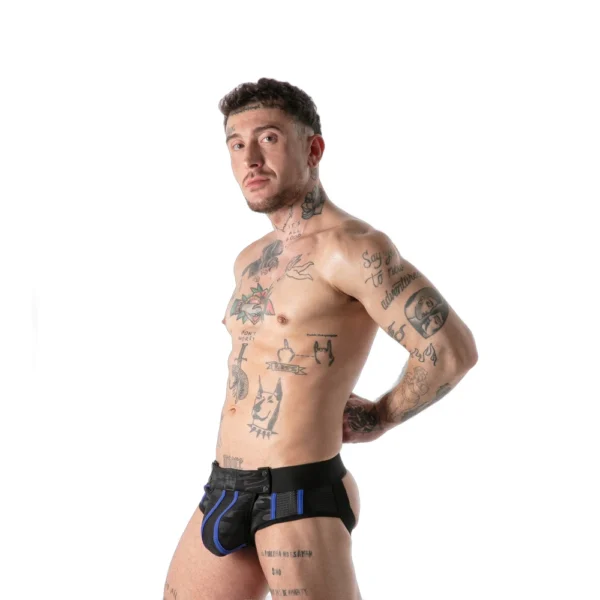 A tattooed man stands in profile against a white background, wearing black and blue harness-style underwear, with his hands resting on his hips and looking towards the camera.