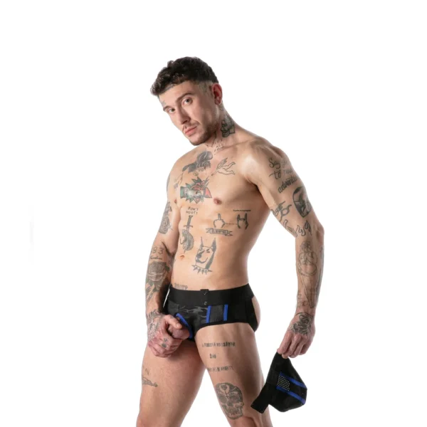 Tattooed man wearing a black jockstrap stands against a white background, holding the front pouch of the underwear in one hand and looking towards the camera.