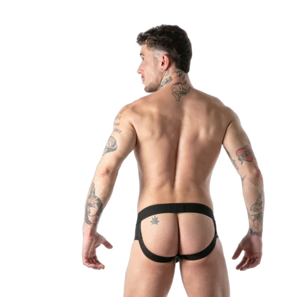 A tattooed man stands facing away, wearing a black jockstrap, showing his bare back and tattoos on arms, shoulders, and thigh, against a white background.
