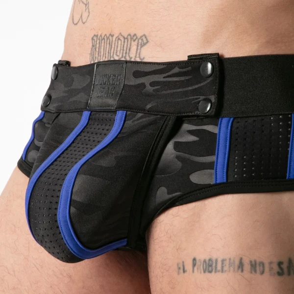A close-up of a person wearing black and blue camouflage pants with mesh details and visible press studs. Tattoos are visible on the person’s lower abdomen and hip.