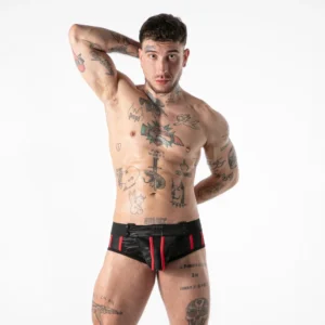 A topless person with tattoos poses with one hand behind their head, wearing black and red shorts against a plain white background.