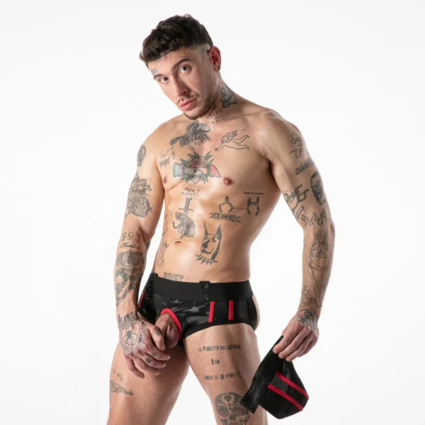 A tattooed man stands against a white background wearing black and red pants with a front opening, holding a matching pouch in his left hand and looking at the camera.