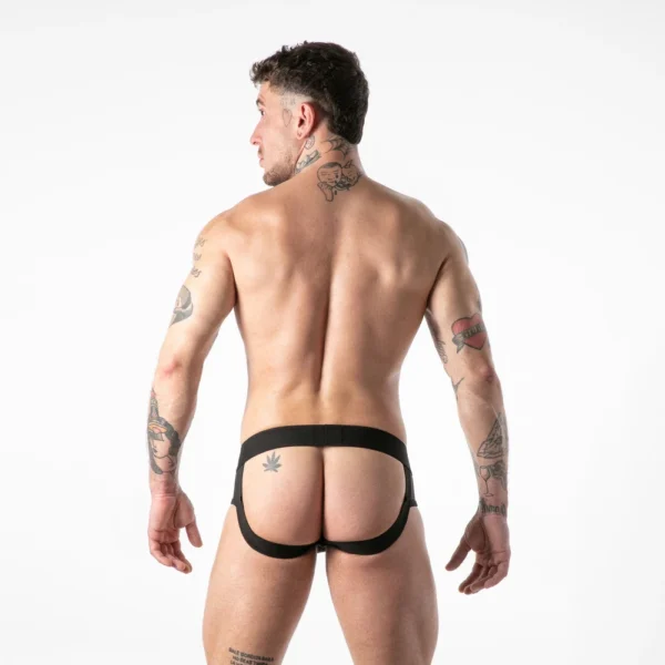 A tattooed man stands with his back to the camera wearing a black jockstrap, exposing his back and legs against a plain white background.