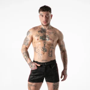 A bare-chested man with tattoos stands against a white background, wearing black shorts with red stripes and one hand in his pocket.