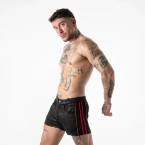 A shirtless man with tattoos stands sideways, looking at the camera. He wears black athletic shorts with red stripes and has short dark hair. The background is plain white.