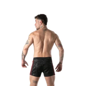 A bare-chested man with tattoos, wearing black camouflage shorts with red stripes, stands facing away against a white background.